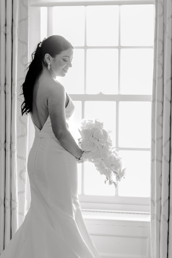 Black and white photo of bride standing in front of window